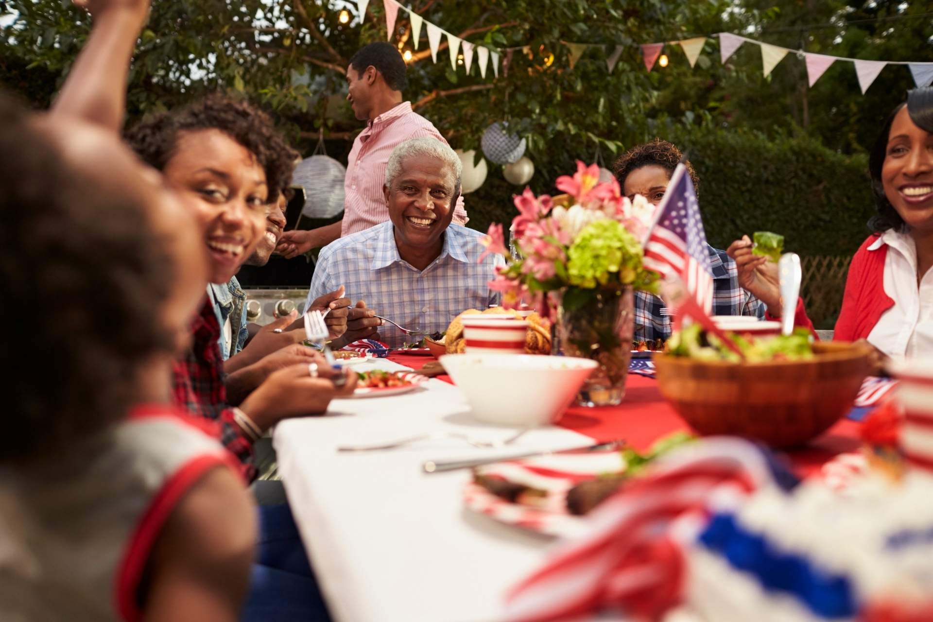 A multigenerational family gathers around an outdoor table decorated with American flags and festive red, white, and blue accents, enjoying a backyard meal together during a Fourth of July celebration.